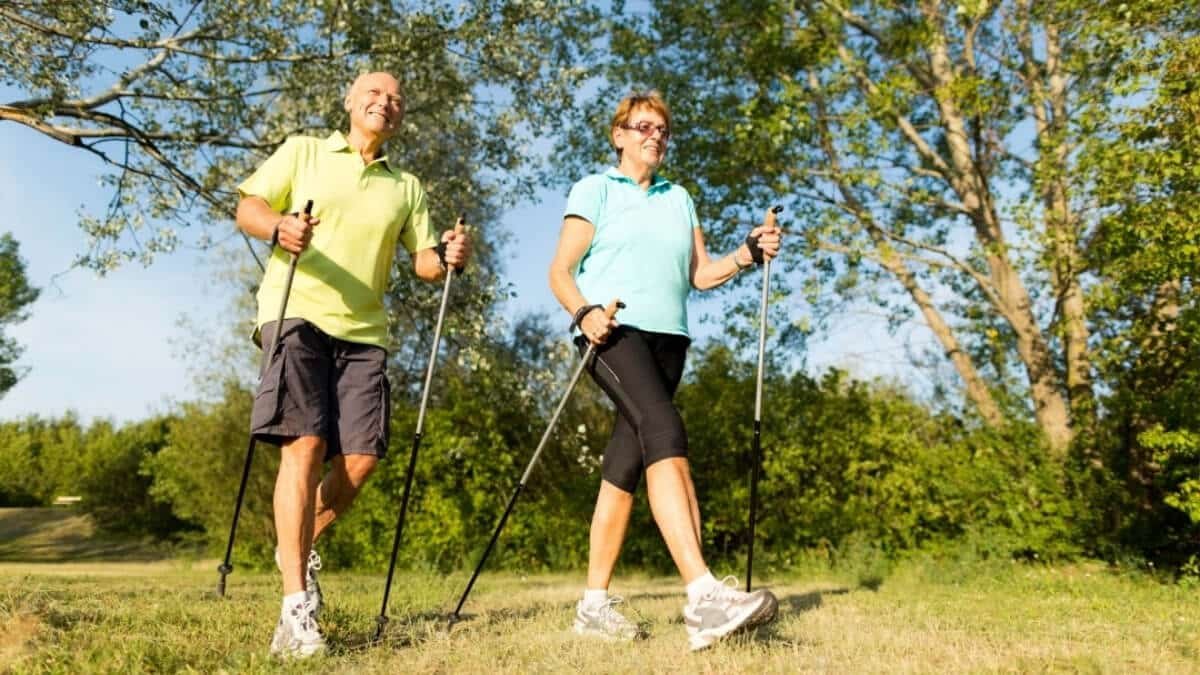 Apžvelgimas šiaurietiškas ėjimas. Paveiksliuke atvaizduot pagyvenusių žmonių pora, vyras ir moteris gamtoje sportuojantis su šiaurietiškomis lazdomis.
