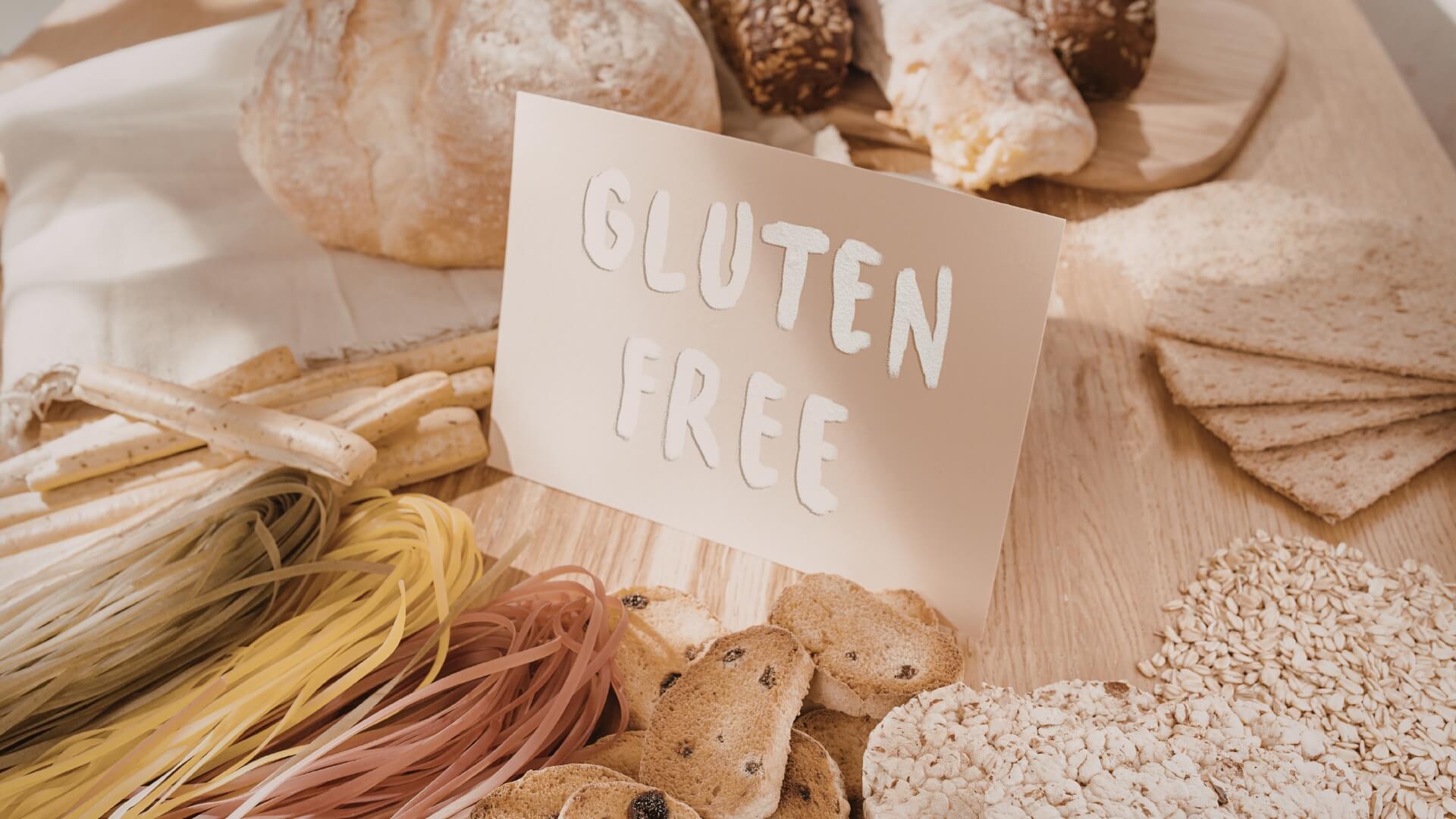 Dieta Be Gliuteno (Gluten-Free Diet) Dieta Be Gliuteno (Gluten-Free Diet). Įvairūs maisto produktai be gliuteno. Makaronai, trapučiai, duona ir grūdai ant stalo, su kortele „gluten free“, pabrėžiančia begliutenės mitybos pasirinkimą.