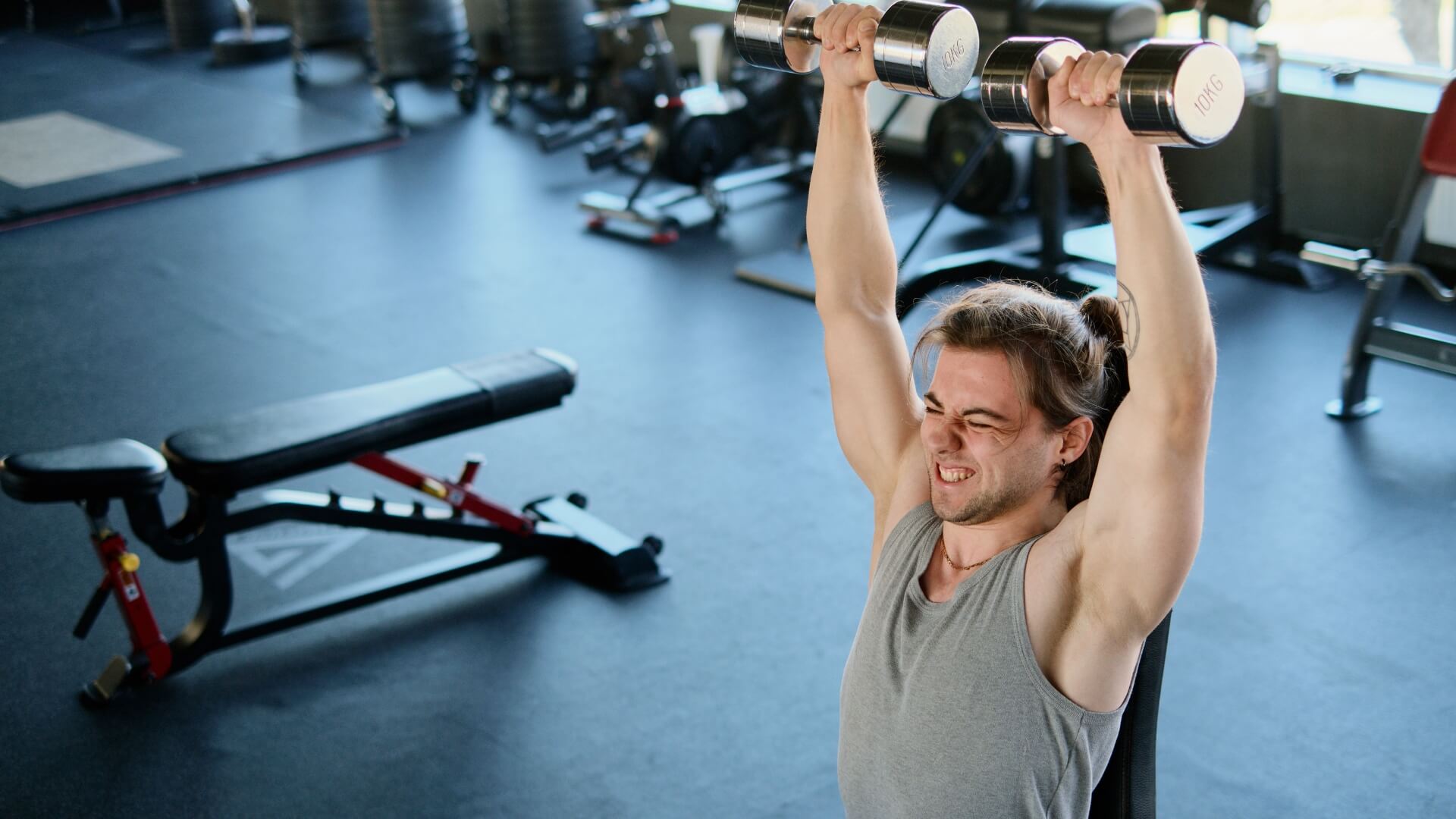Kiek Kalorijų Reikia Raumenų Auginimui (How Many Calories for Muscle Growth). Kiek Kalorijų Reikia Raumenų Auginimui (How Many Calories for Muscle Growth). Sporto salėje vyras kelia hantelius virš galvos, iliustruojant jėgos treniruotes, kurios kartu su tinkamu kalorijų kiekiu skatina raumenų augimą.