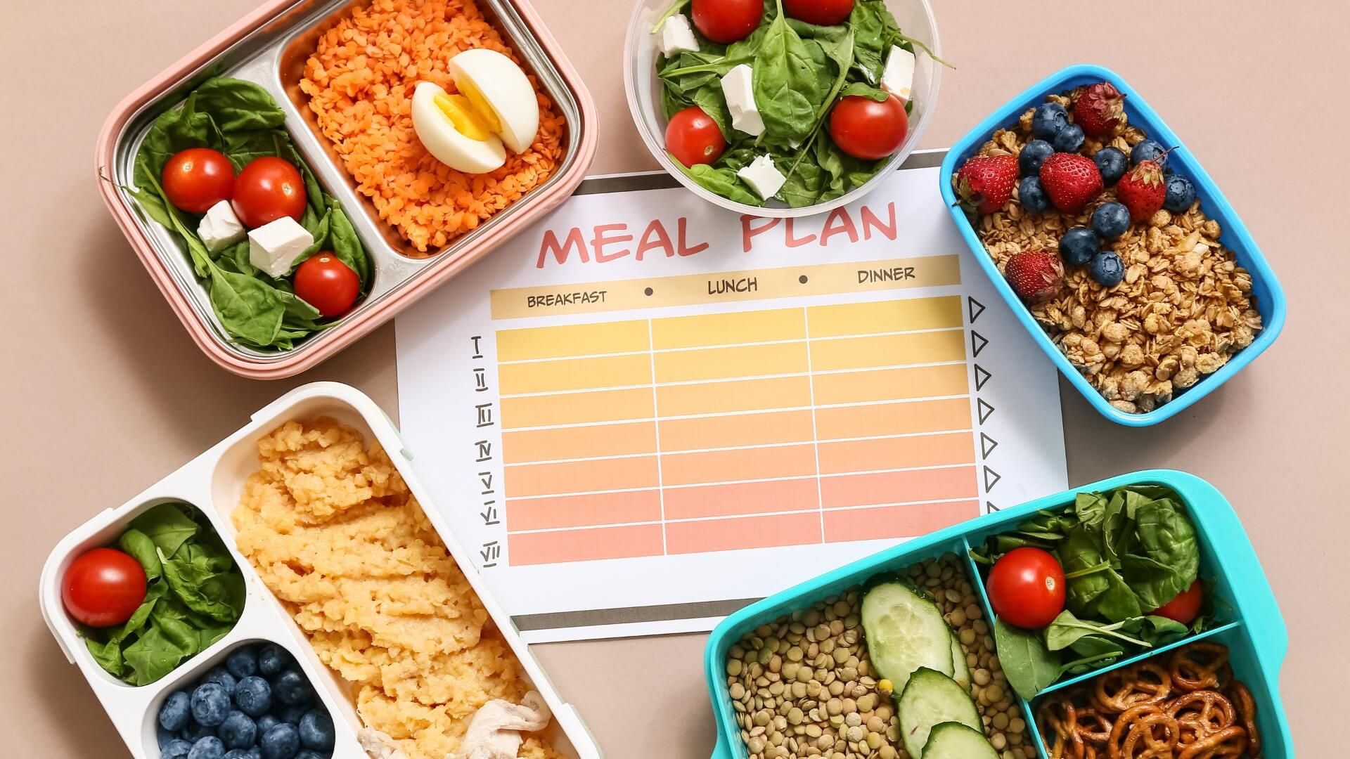 Komerciniai Mitybos Planai (Commercial Diet Plans) Komerciniai Mitybos Planai (Commercial Diet Plans). Paveikslėlyje pavaizduota savaitės mitybos plano lentelė, apsupta įvairių porcijuotų maisto dėžučių su subalansuotais produktais (daržovės, kruopos, vaisiai, baltymai), iliustruojant komercinių mitybos planų struktūruotą, iš anksto suplanuotą ir porcijomis kontroliuojamą mitybą.