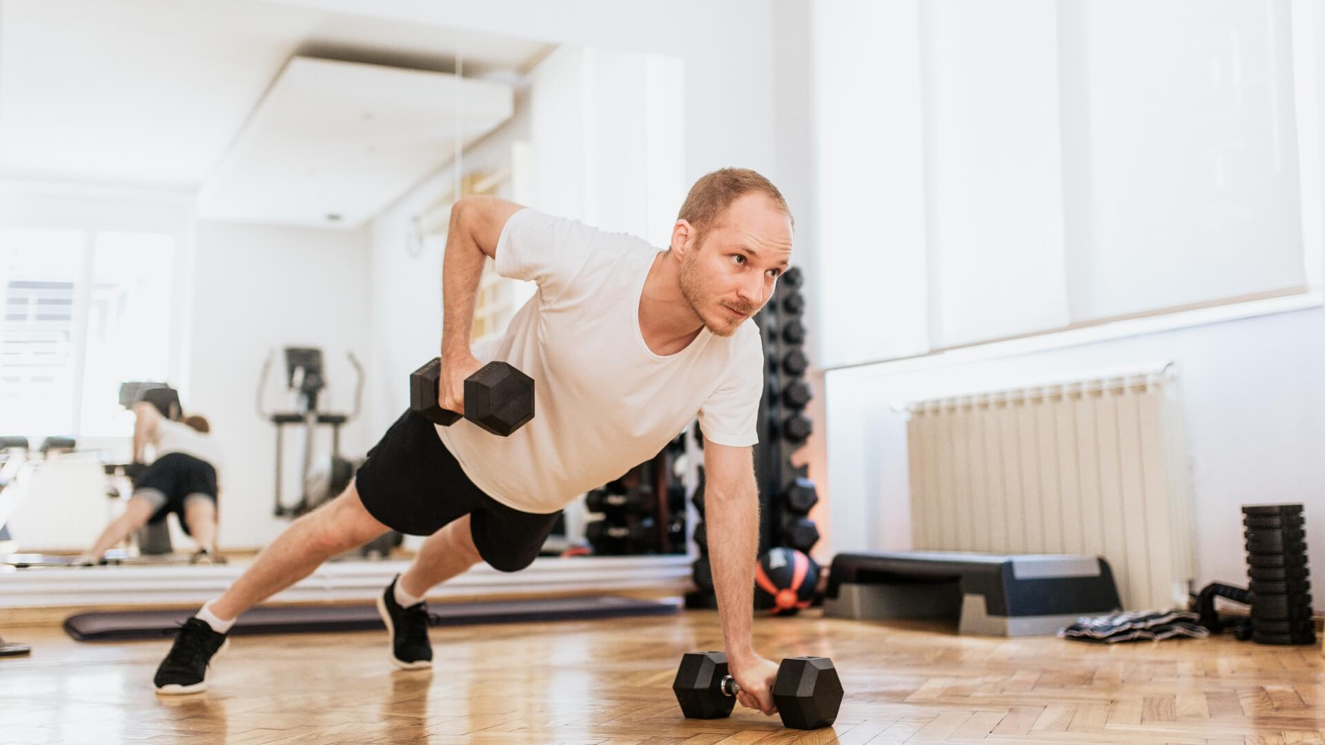 Koks Kartojimo Tempas Geriausias Raumenų Augimui? Koks Kartojimo Tempas Geriausias Raumenų Augimui?. Vyras sporto salėje atlieka kombinuotą jėgos pratimą su hanteliais – atsispaudimą su traukimu (renegade row), lavindamas viršutinės kūno dalies raumenis, stabilumą ir koordinaciją.