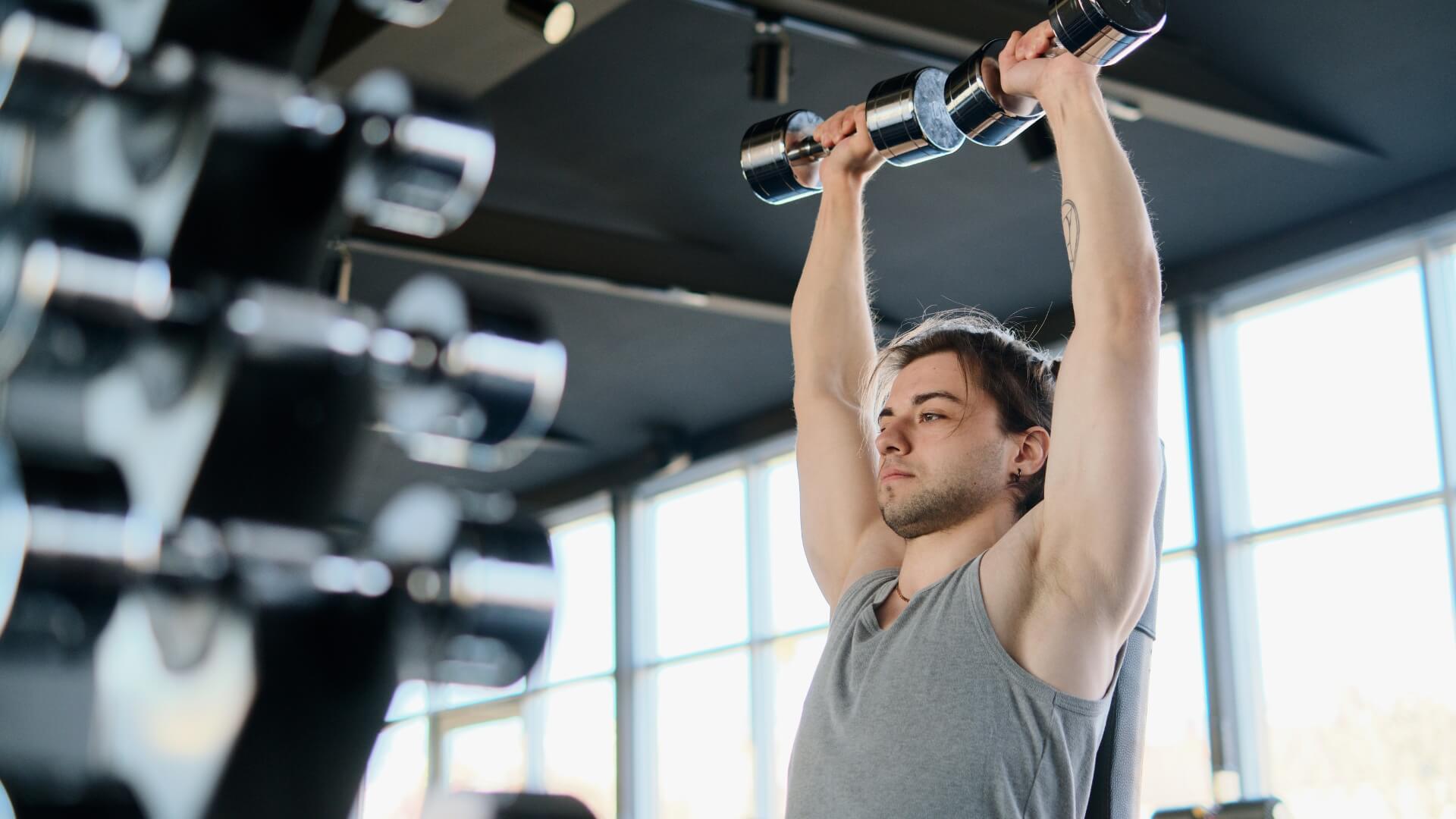 Koks Kartojimo Tempas Geriausias Raumenų Augimui? Koks Kartojimo Tempas Geriausias Raumenų Augimui?. Vyras sporto salėje atlieka hantelių spaudimą virš galvos sėdėdamas, lavindamas pečių raumenis ir viršutinės kūno dalies jėgą.