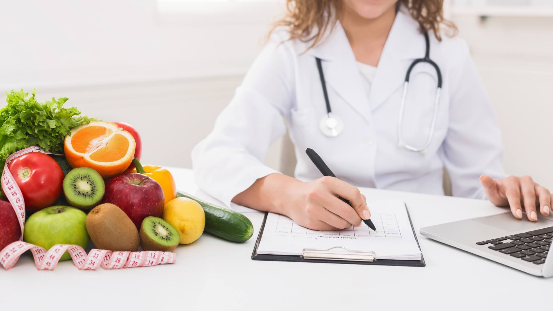 Kraujo Grupės Dieta (Blood Type Diet) Kraujo Grupės Dieta (Blood Type Diet). Dietologas rašo mitybos planą prie stalo, šalia padėti vaisiai, daržovės ir matavimo juosta.
