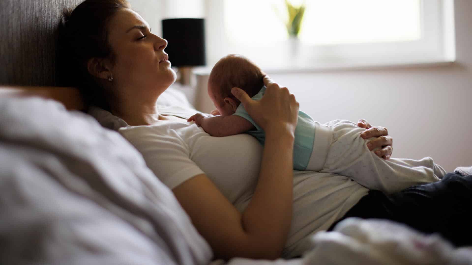 Pogimdyminis Terapinis Masažas – Masažo Studija „Humanus“. Nuotraukoje mama ilsisi lovoje, švelniai priglaudusi naujagimį prie krūtinės. Jauki motinos ir kūdikio akimirka, perteikianti rūpestį, poilsį ir dėmesį mamos savijautai po gimdymo.
